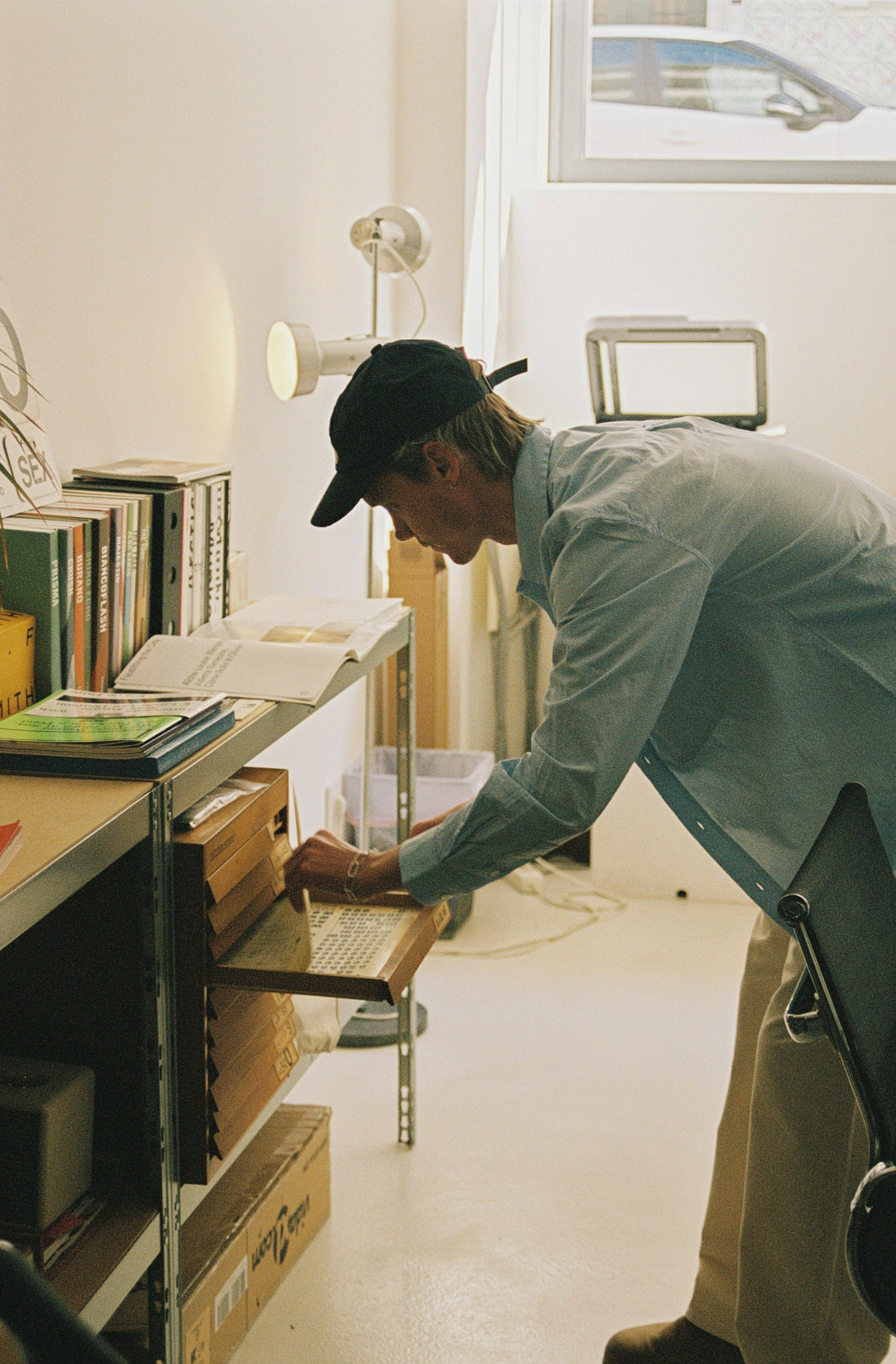 A man in a light blue shirt, khaki pants, and a black baseball cap leans over a metal shelving unit in a bright, minimalist room, reaching for a wooden drawer.