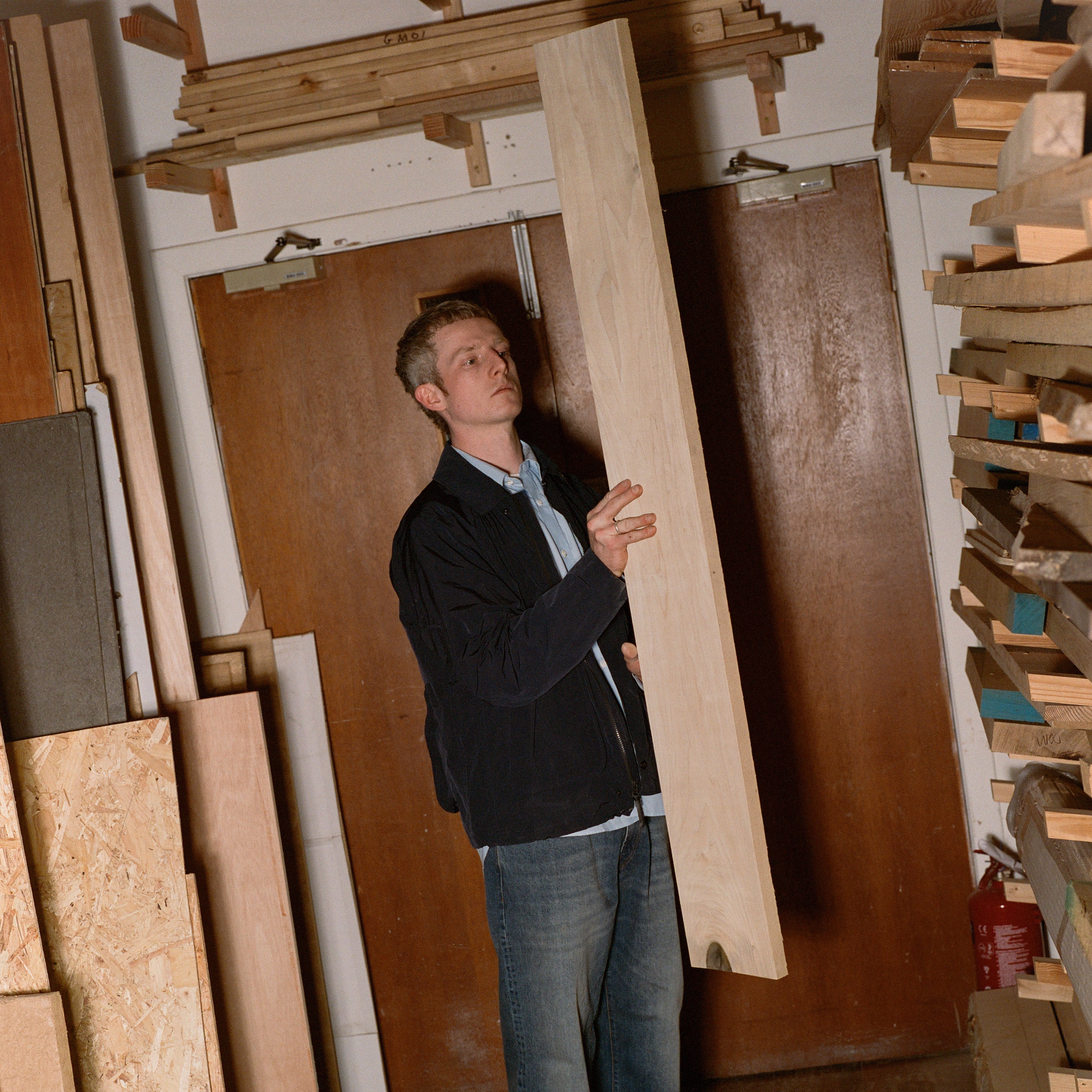 Man holding a large wooden board in a workshop filled with wood planks.