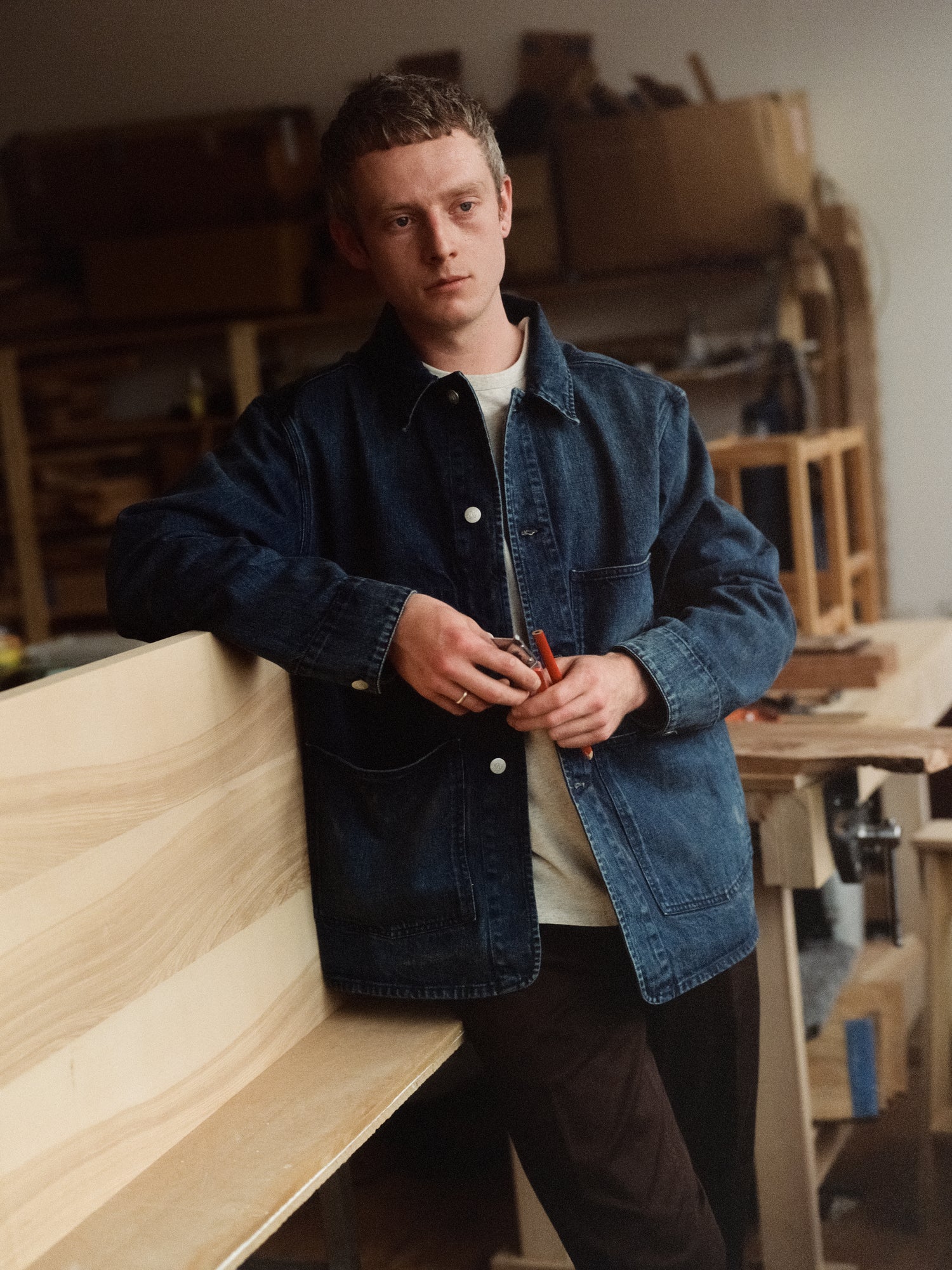 Man in a workshop holding a tool, wearing a blue denim jacket.